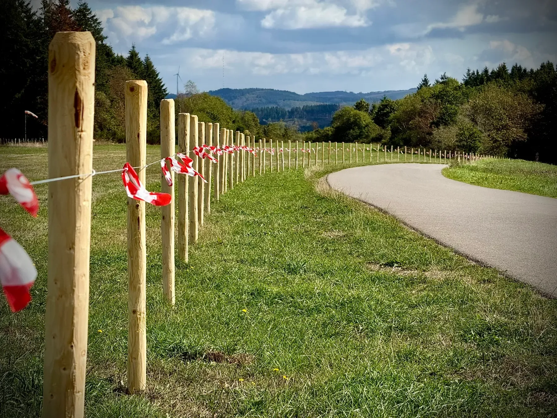 Piquets en bois reliés par une rubalise rouge et blanche, marquant la limite de la zone de chantier le long du chemin goudronné