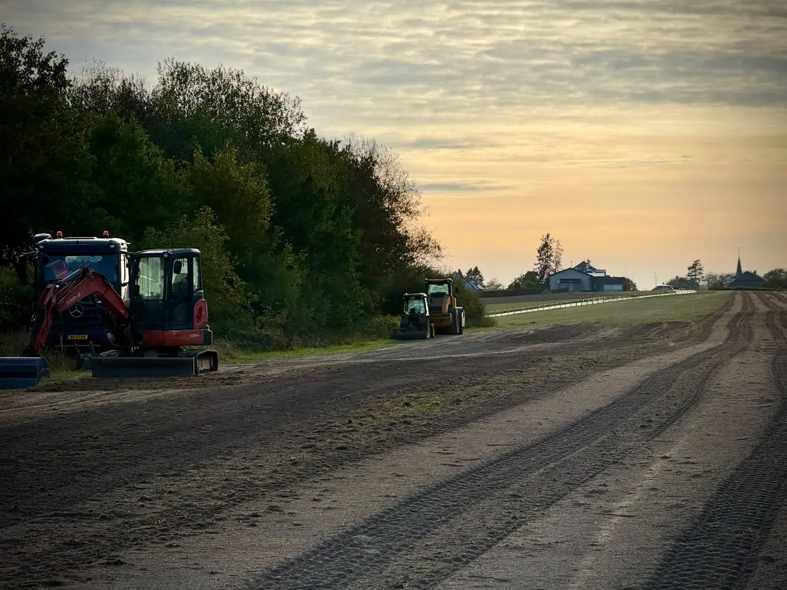 Entlang der Baumreihe neben der Piste abgestellte Maschinen in der Abenddämmerung, am Horizont das Dorf mit seinem Kirchturm