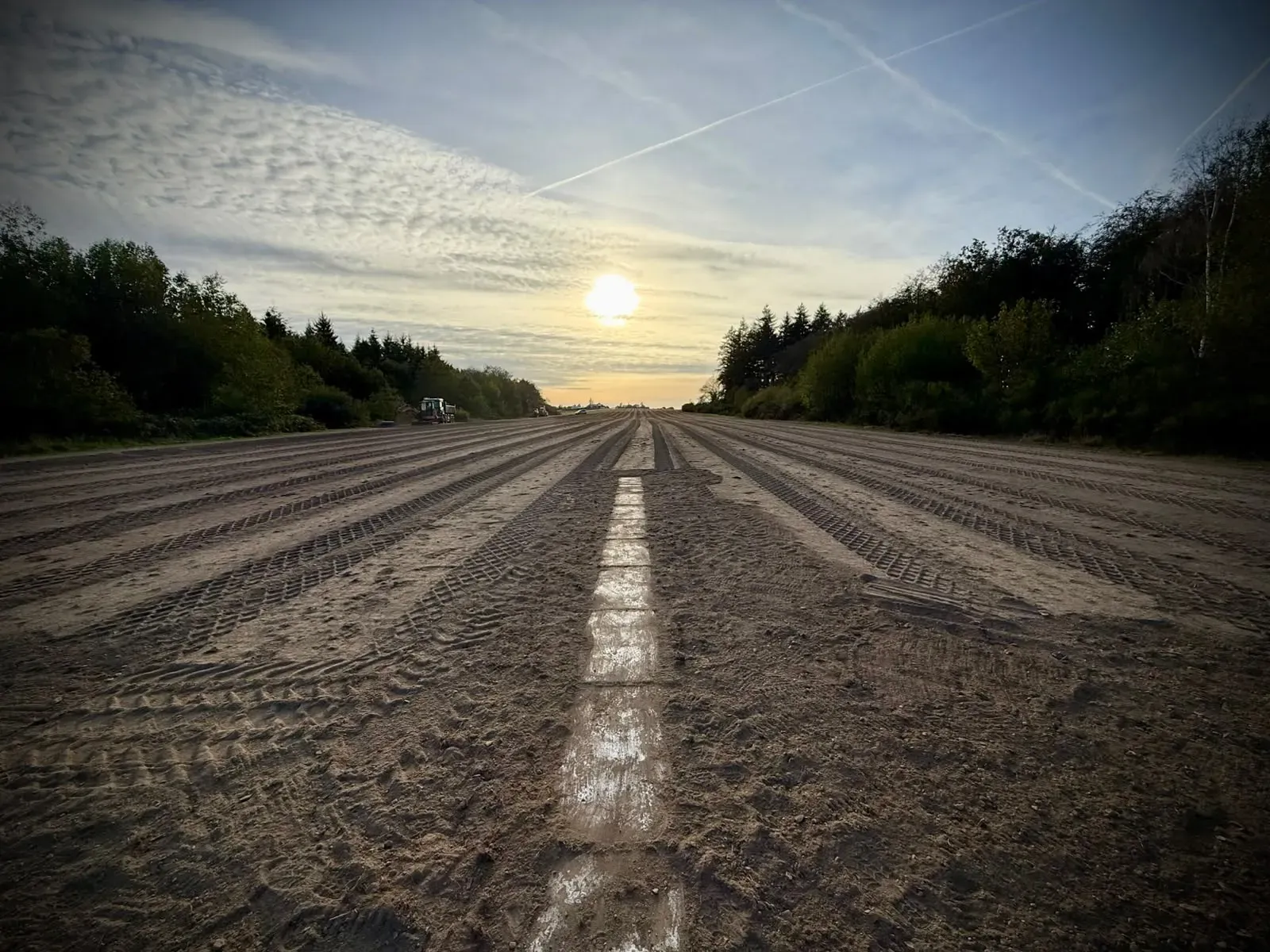 Die Piste aus blankem Erdreich bei Sonnenuntergang, Blick entlang einer aufgemalten Mittellinienmarkierung, am anderen Ende Maschinen zwischen den Bäumen