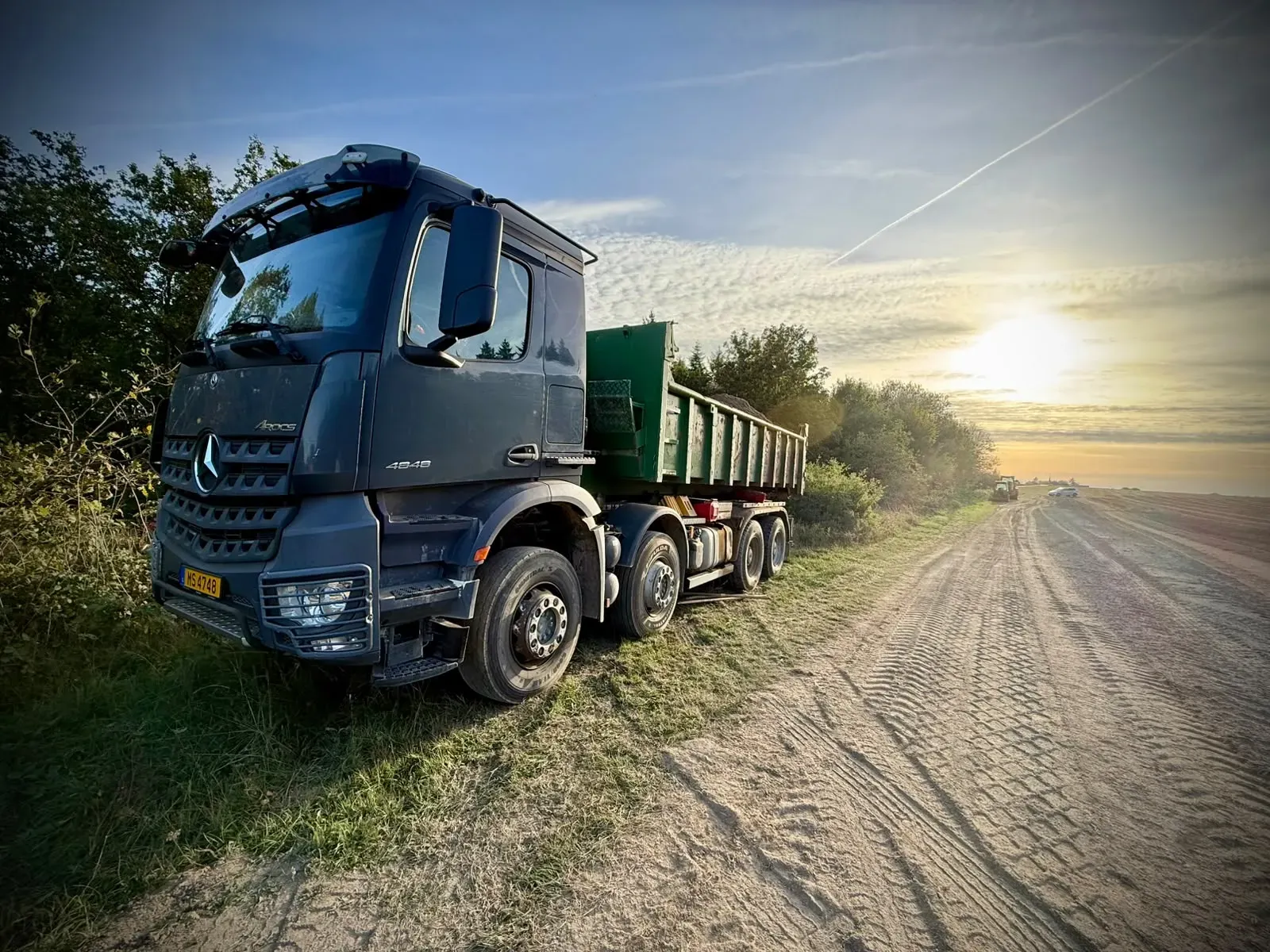 Ein schwerer Lastwagen mit beladenem Container, auf der Grasfläche neben der Piste abgestellt, dahinter die untergehende Sonne