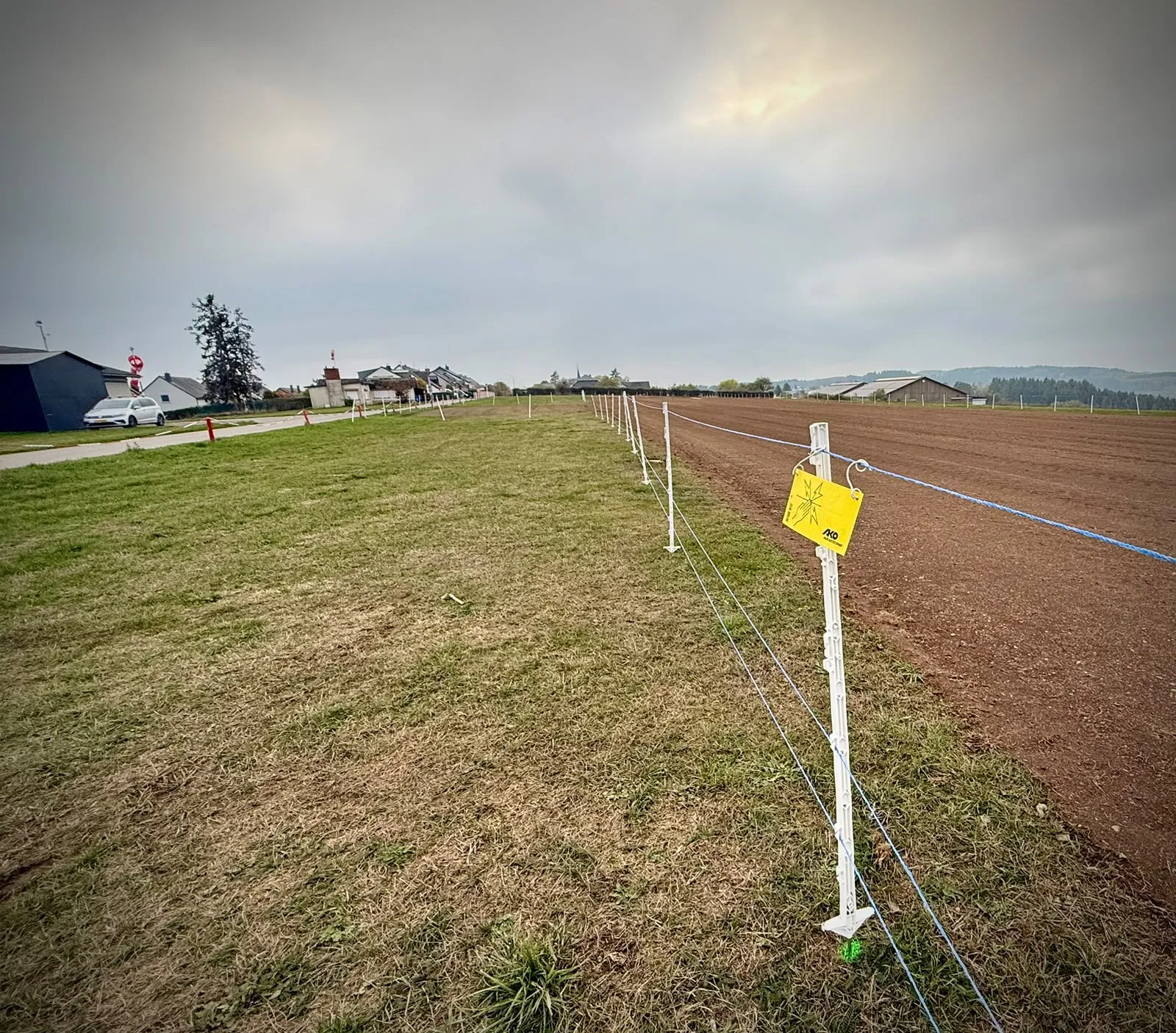 Piquets et fils d'une clôture électrique avec un panneau d'avertissement jaune à côté de la piste, de l'herbe à gauche et le sol préparé à droite