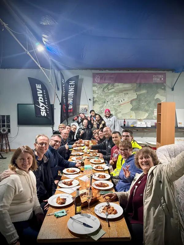 Eine Gruppe lächelt an einer langen gedeckten Tafel in der Vereinshalle zur Kamera, mit Tellern, Gläsern und Flaggen im Bild